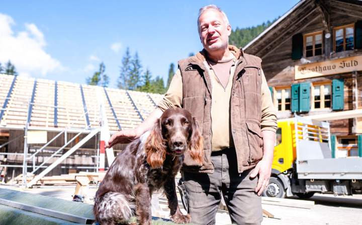 Adrian Bircher mit seinem Hund Quna vor der «Suld-Beiz» und dem Sägereigebäude (links), das als erstes mit Photovoltaik ausgerüstet wird. BILD: HSF