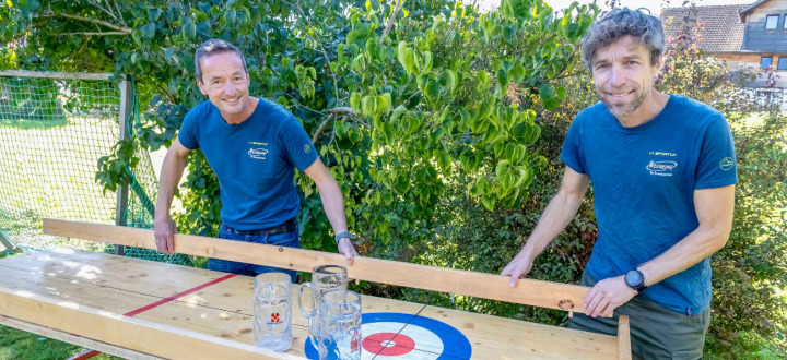 Jürg Schranz (l.) und Daniel Wandfluh zeigen, wie das Bierhumpen-Curling funktioniert: Wer zirkelt den Bierhumpen über eine fünf Meter lange Bahn direkt in die Mitte des Curling-Kreises? BILD: ZVG